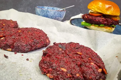 Gotowe burgery z buraka na papierze do pieczenia, jeden złożony burger z bułką i dodatkami w tle.