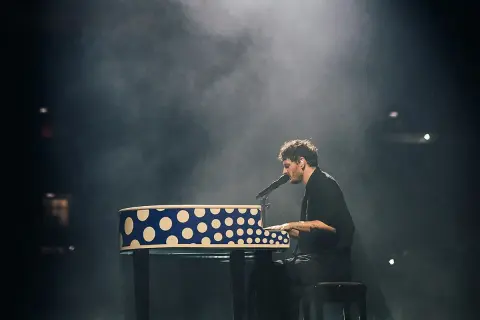 Dawid Podsiadło na koncercie, grając na pianinie w niebiesko-białe kropki, otoczony mgłą sceniczną.