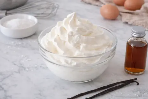 Un bol de crème chantilly aérienne, prêt à être dégusté. À côté, des gousses de vanille et une petite bouteille d'extrait.