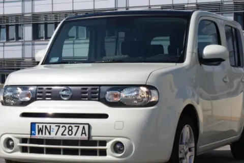 Biały Nissan Cube z polskimi tablicami rejestracyjnymi WN 7287A zaparkowany przed nowoczesnym budynkiem.