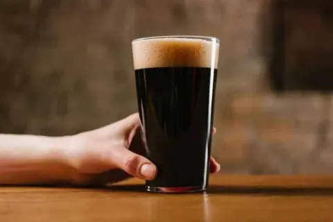 Ręka trzymająca szklankę ciemnego piwa porter. Czy piwo porter jest zdrowe? To pytanie zadaje sobie wielu miłośników tego trunku.