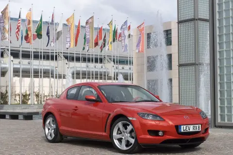 Czerwona Mazda RX-8 zaparkowana przed budynkiem z fontannami i flagami.