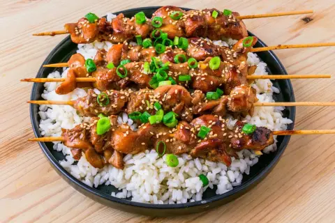 Fyra kycklingspett teriyaki ligger på en bädd av vitt ris, toppade med sesamfrön och hackad salladslök.