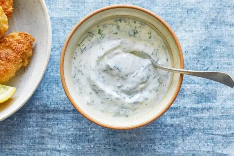 En skål med krämig kall sås till fisk, med dill och en sked. Bredvid syns friterad fisk och citronklyftor.