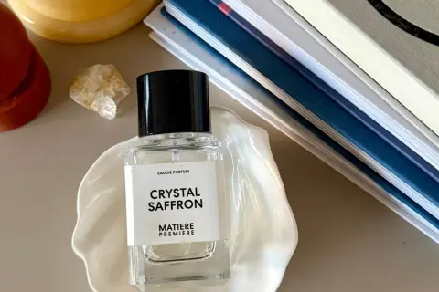 Elegancka butelka perfum "Crystal Saffron" na muszli, obok świecy i książek. Zapach orientalny, który przeniesie Cię w podróż.