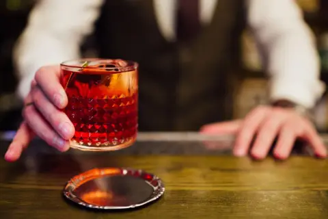 Ein Barkeeper reicht einen roten Cocktail mit Eis und Garnitur. Perfekt, um cocktail mixen lernen.
