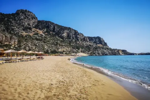 Piękna plaża Tsambika z piaszczystym brzegiem, turkusową wodą i skalistym zboczem w tle. Leżaki i parasole zapraszają do odpoczynku.