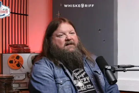 A man with a long beard and hair, wearing a denim jacket, speaks into a microphone. He's discussing Channing Wilson songs, possibly at a music festival.