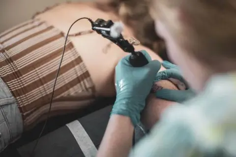 Tatuador con guantes azules haciendo un tatuaje de puñal en la pierna de una persona.