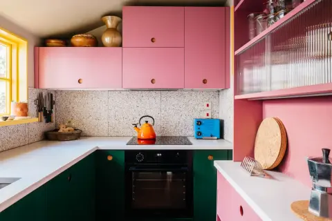 Cucina rosa cipria con mobili verdi, piano cottura, tostapane blu e decorazioni in terracotta.