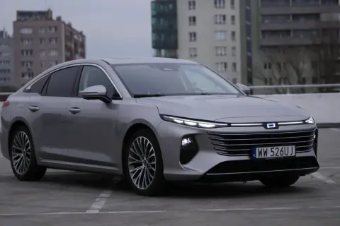 Srebrny Bestune B70 z nowoczesnymi światłami LED zaparkowany na betonowym placu, w tle budynki mieszkalne.