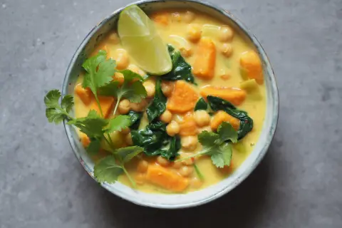 Aksamitne curry z ciecierzycy z kawałkami batata i szpinaku, ozdobione świeżą kolendrą i ćwiartką limonki.