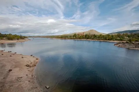 El embalse del Pontón, con aguas azules y orillas arenosas, se extiende hacia colinas verdes y un edificio de ladrillo rojo bajo un cielo nublado.