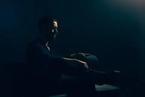 Mężczyzna z brodą siedzi w półmroku, wpatrzony w dal. Jego postawa jest **low key**, emanuje spokojem i zadumą.