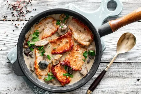 Pyszny schab w sosie grzybowym z natką pietruszki, podany w patelni na drewnianym blacie. Obok leży stara łyżka.