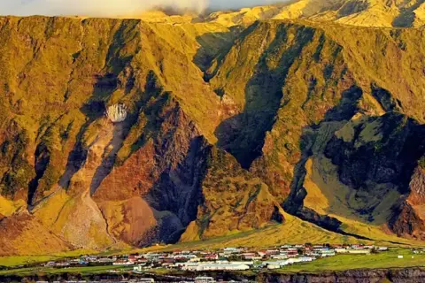 Un pueblo en Tristan de Acuña se asienta al pie de imponentes montañas cubiertas de hierba, bañadas por la luz dorada del atardecer.