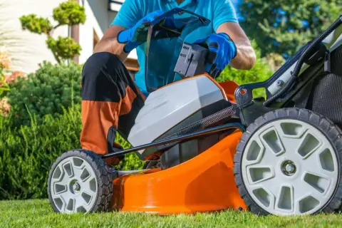 Mężczyzna wymienia baterię w pomarańczowej kosiarce akumulatorowej, przygotowując się do pracy w ogrodzie. To ważny element w rankingu kosiarek akumulatorowych.