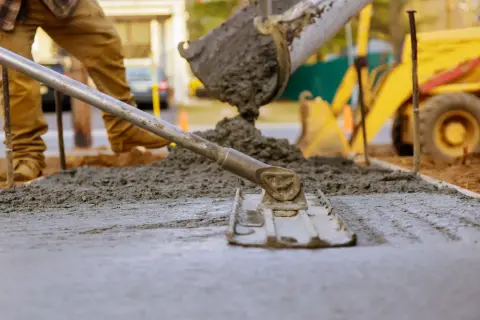 Pracownik wylewa beton na chudziaka, używając łaty do wyrównania powierzchni. W tle widać koparkę.
