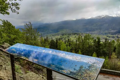 Widok z wieży widokowej Karkonosze. Na pierwszym planie tablica informacyjna z mapą panoramy, w tle góry i lasy.