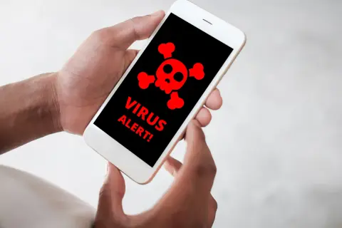 Dłonie trzymające smartfon z czerwonym symbolem czaszki i napisami "VIRUS ALERT!".
