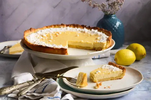 Eine köstliche Tarte au Citron, bestäubt mit Puderzucker, steht bereit. Ein Stück wurde bereits auf einem Teller serviert, daneben liegen frische Zitronen.