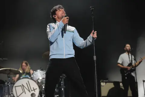 Louis Tomlinson na scenie, śpiewający do mikrofonu. W tle perkusista i gitarzysta.