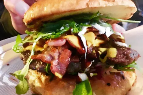 Soczysty burger z bekonem, serem, rukolą i sosami, trzymany w dłoni.