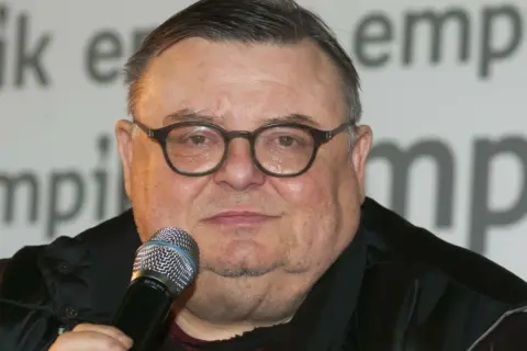 Wojciech Mann, znany z audycji radiowych, w okularach i z mikrofonem. Jego wiek nie jest widoczny na zdjęciu.