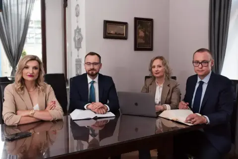 Cztery osoby w strojach biznesowych siedzą przy stole w sali konferencyjnej, przed nimi dokumenty i laptop.