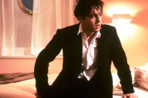 Hugh Grant w garniturze, w rozpiętej koszuli, wygląda na zamyślonego. Scena z jednego z jego filmów.