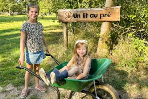 Deux petites filles profitent d'un séjour à la ferme, l'une assise dans une brouette, l'autre à côté, sous un panneau "Un lit au pré".