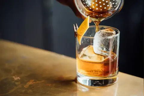 Bursztynowy drink z lodem i skórką pomarańczy, nalewany z shakera. Miękkie, rozmyte tło tworzy przyjemny **bokeh**.