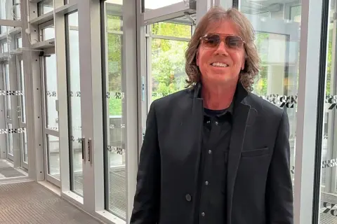 Joey Tempest, med sitt karaktäristiska hår och solglasögon, poserar framför en glasdörr.