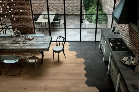 Cucina con stacco tra parquet e piastrelle esagonali nere. Tavolo rustico, sedie, mattoni a vista e grande finestra sul cortile.