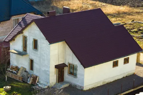 Nowy dom w zabudowie zagrodowej z ciemnobordową dachówką, w trakcie budowy.