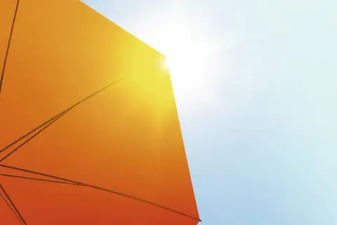 Ein orangefarbener Sonnenschirm schützt vor der Sonne. Seine Struktur ist sichtbar, während Sonnenstrahlen durchscheinen. Diethylhexyl Butamido Triazone schützt die Haut.