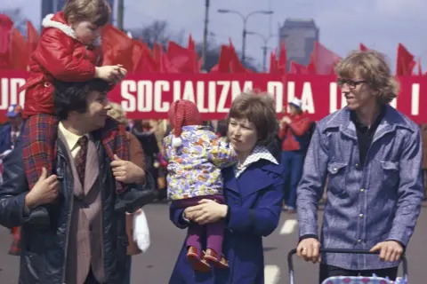 Rodzina na tle czerwonego transparentu z napisem "SOCJALIZM DLA PRACY" i czerwonych flag.