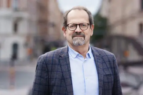 Peder Hasslev, en man med glasögon och skägg, bär en rutig kavaj och ljusblå skjorta. Bakgrunden är en suddig stadsmiljö.
