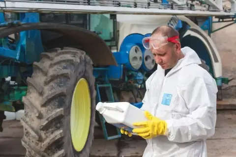 Rolnik w kombinezonie ochronnym i rękawicach trzyma butelkę z preparatem, prawdopodobnie zawierającym difenokonazol, obok traktora.
