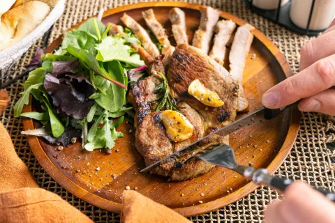 Ein saftiges Schweinesteak in der Pfanne, mit Rosmarin und Knoblauch verfeinert, wird mit einem Messer und einer Gabel angeschnitten. Dazu gibt es einen frischen Salat.