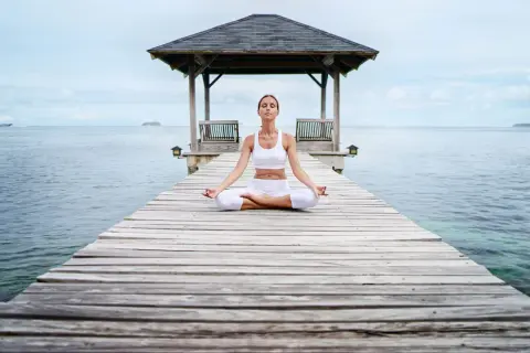Frau in weißer Kleidung meditiert auf einem Steg am Meer. Eine Anleitung zur transzendentalen Meditation, um inneren Frieden zu finden.