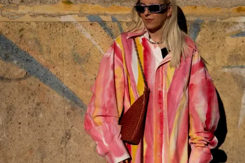 Kobieta w okularach przeciwsłonecznych i koszuli w stylu tie-dye w odcieniach różu i żółci, z brązową torebką na łańcuszku.