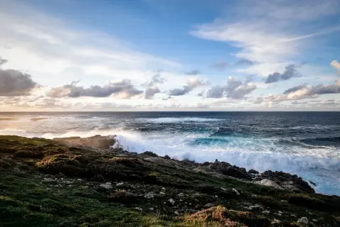 O mar bravo bate nas rochas rochosas, espumando e rugindo. O céu azul com nuvens brancas paira sobre o vasto oceano, um cenário perfeito para o portiño.