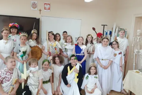 Dzieci w strojach antycznych bogów i bogiń, jak Artemida, pozują do zdjęcia. W tle szkielet i tablica.