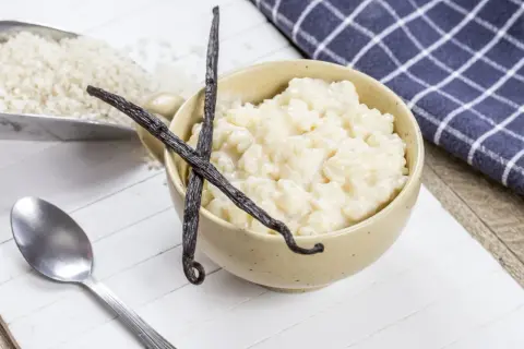 Un bol de riz au lait crémeux, garni de gousses de vanille, avec du riz brut dans une pelle à côté.