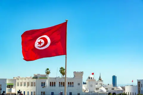 Tunisiens flagga vajar stolt mot en klarblå himmel över vita byggnader. En påminnelse om en resa till Tunisien.