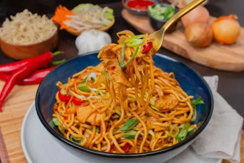 Bami Goreng mit Hühnchen, garniert mit Frühlingszwiebeln und Chili. Daneben sind Zutaten wie roter Chili, Knoblauch und Zwiebeln zu sehen.