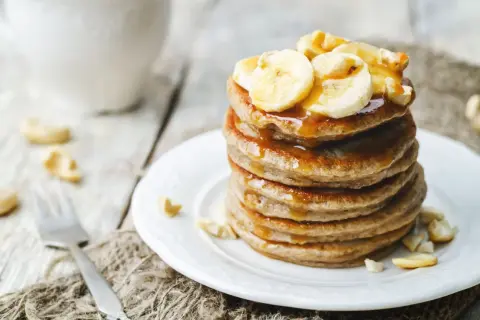 Ein Stapel goldbrauner Bananenpfannkuchen, beträufelt mit Sirup und garniert mit Bananenscheiben und Cashewkernen.
