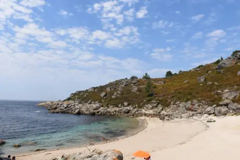 Playa rocosa y arena dorada en **Cabo Udra**, con aguas cristalinas y un cielo azul con nubes.