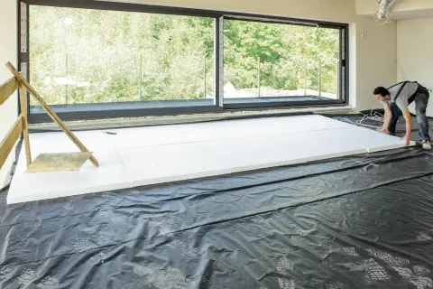 Pracownik układa płyty styropianowe na folii, przygotowując się do tego, jak ocieplić strop betonowy styropianem. Duże okno wychodzi na zieleń.